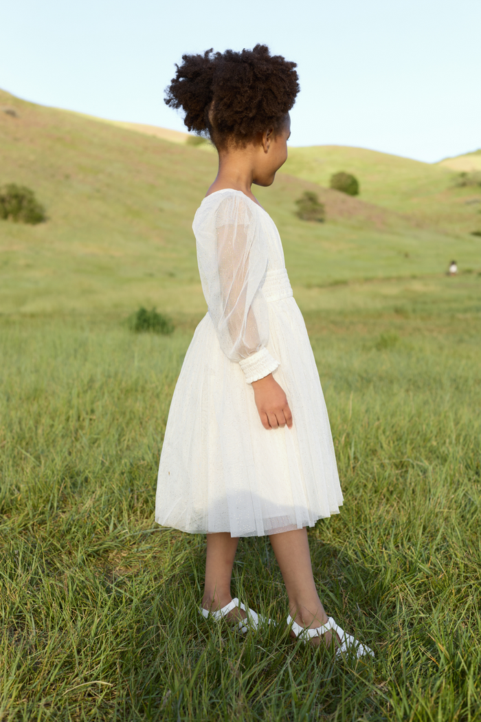 A young girl wearing the Ivy City Co Mini Stella Dress in White and matching white shoes stands on green grass, gazing at rolling hills under a clear sky.