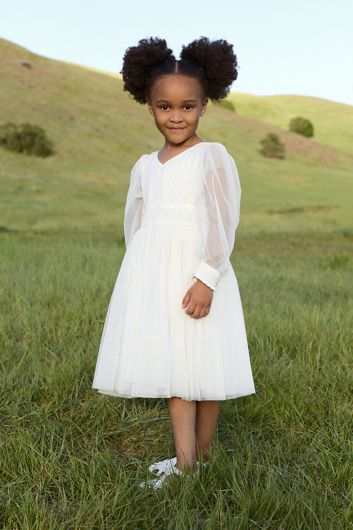 A young flower girl, dressed in the Ivy City Co Mini Stella Dress in White, stands smiling on green grass with rolling hills behind her on a clear day.