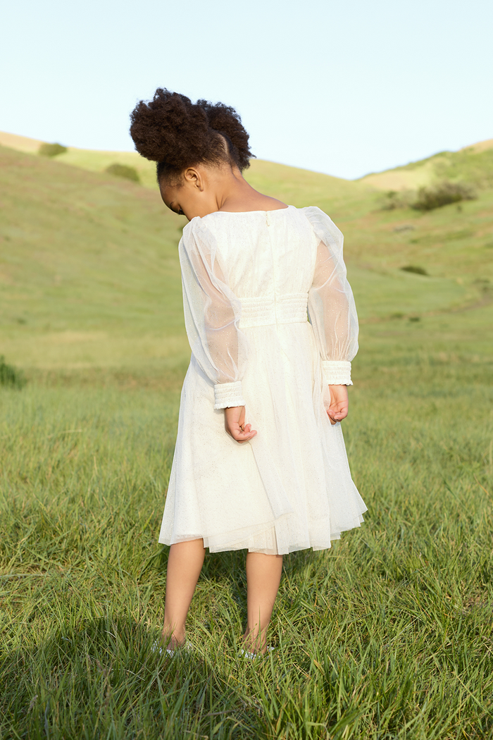 A young girl in the Ivy City Co Mini Stella Dress in White stands barefoot on green grass, her face turned away as she gazes downward, surrounded by rolling grassy hills beneath a clear sky.