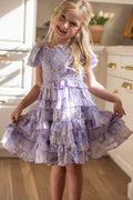 A young blonde girl smiles and holds out the sides of her Ivy City Co Mini Winnie Dress in Lavender Aquarelle Fleur while standing in a bright kitchen with white cabinets and a potted plant visible in the background.