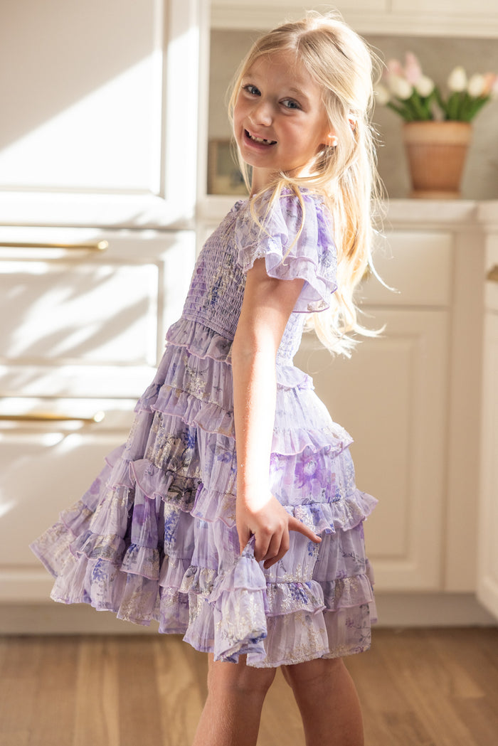A young blonde girl smiles and twirls in the Ivy City Co Mini Winnie Dress in Lavender Aquarelle Fleur, featuring light purple florals, in a bright kitchen with white cabinets and a potted plant.