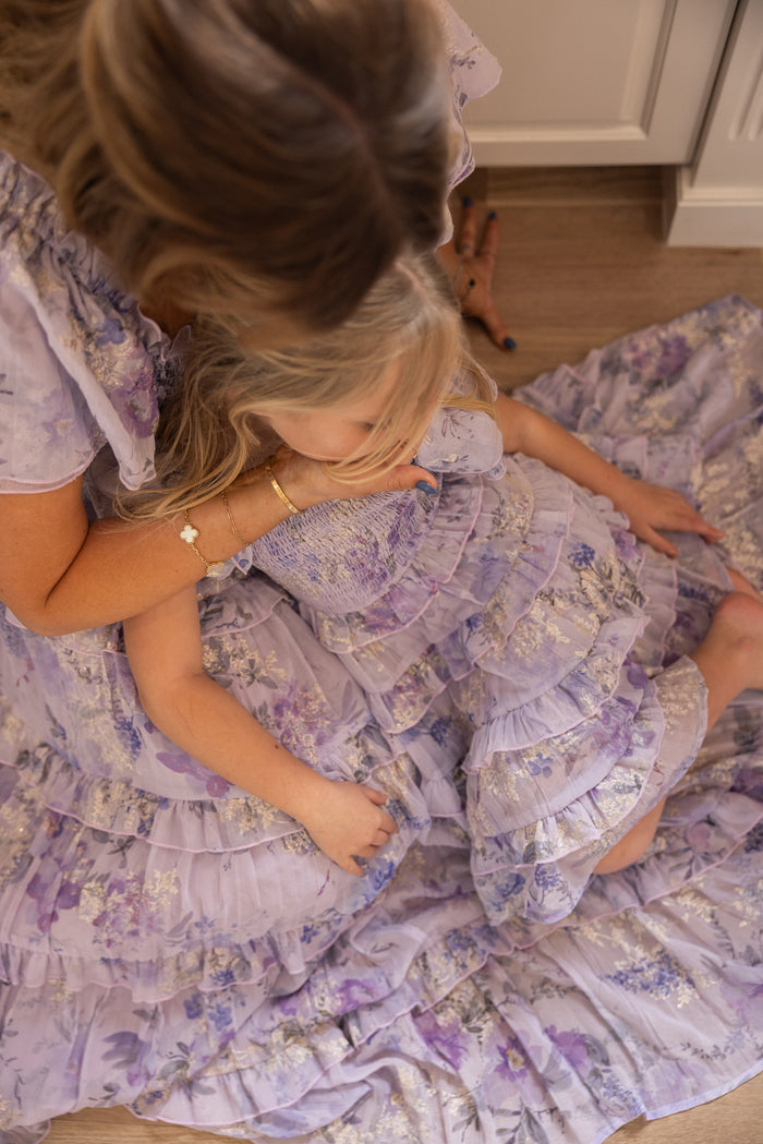 A woman and a young girl sit on the floor near a white cabinet, sharing a cozy moment in matching Ivy City Co Mini Winnie Dresses in Lavender Aquarelle Fleur, their blonde hair glowing against the wooden floor.