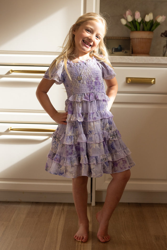 A young girl with long blonde hair stands barefoot on a wooden floor, smiling and posing in the Mini Winnie Dress in Lavender Aquarelle Fleur by Ivy City Co, set against a bright kitchen with white cabinets and a vase of tulips.
