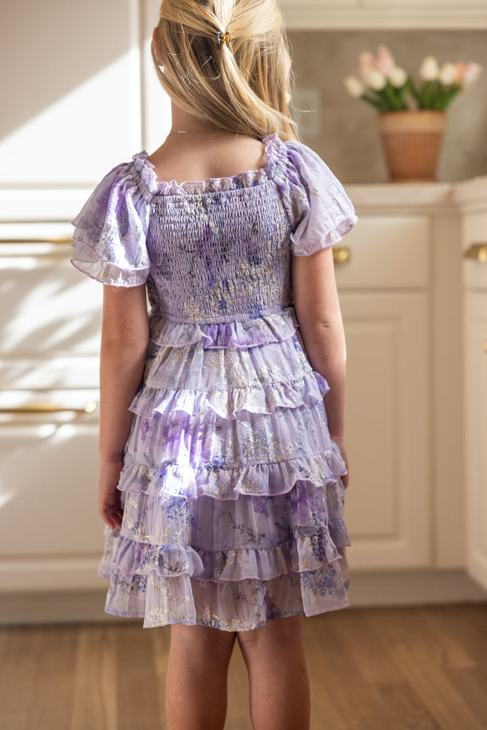 A young girl with blonde hair stands in a kitchen with white cabinets and a potted flower plant, wearing the Ivy City Co Mini Winnie Dress in Lavender Aquarelle Fleur. She is facing away from the camera.