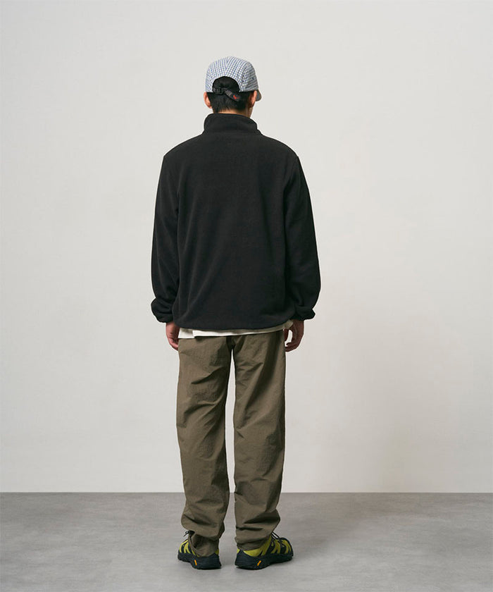 Back view of a model wearing burnt olive colored Nylon Gramicci Pants and a black jacket on a light-tone background.