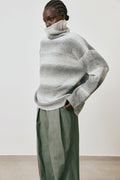Person wearing a large, loose-fitting gray sweater with green pants against a plain background