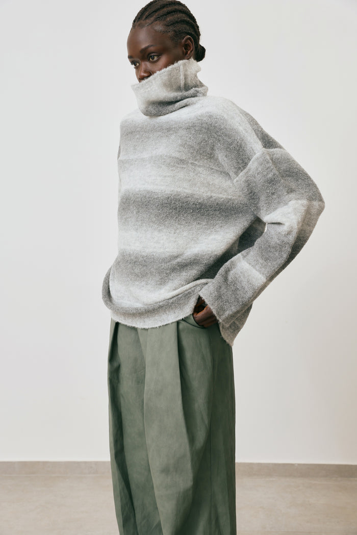 Person wearing a large, loose-fitting gray sweater with green pants against a plain background