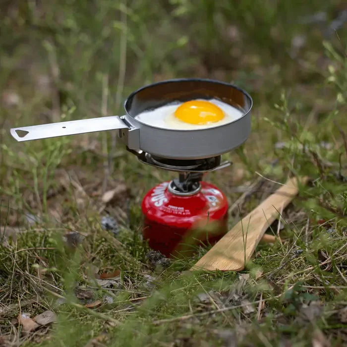 Primus Essential Trail Camping Gas Stove used with a Trek pan lid to fry an egg in a grassy outdoor setting
