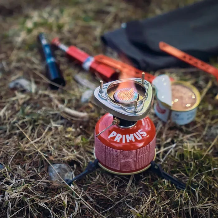 Primus Lite Plus backpacking stove lit on a grassy ground with camping gear in the background