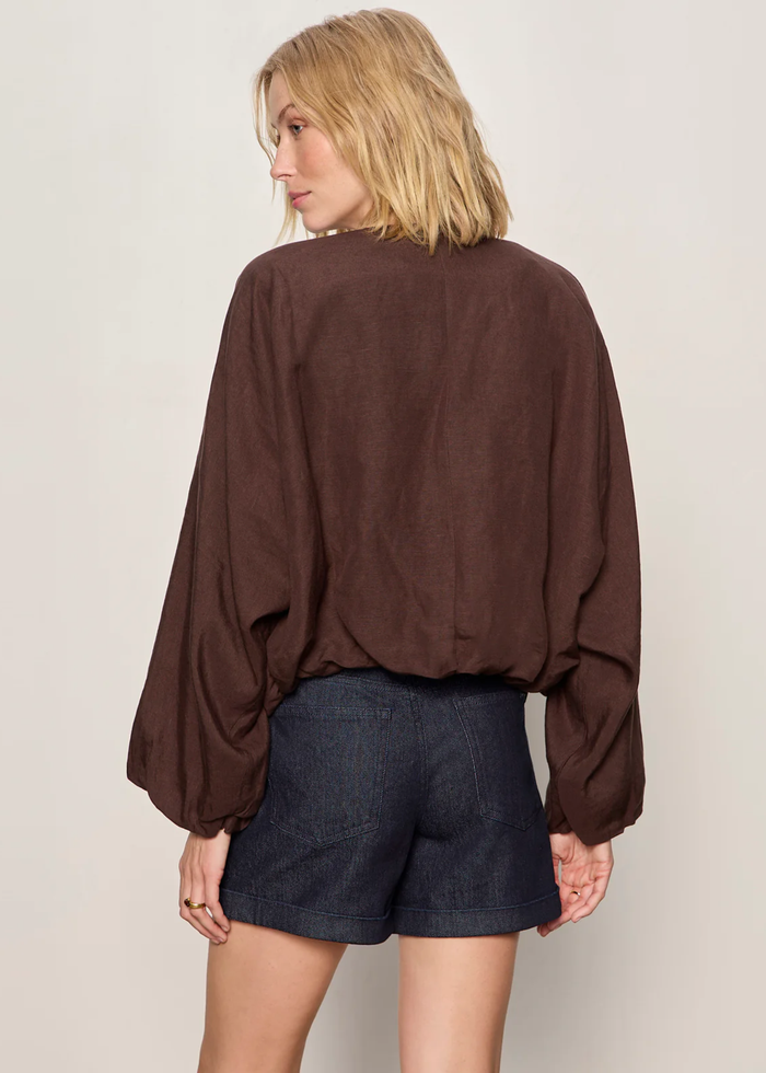 Woman turned around wearing a brown linen jacket with a cream tank and dark blue shorts