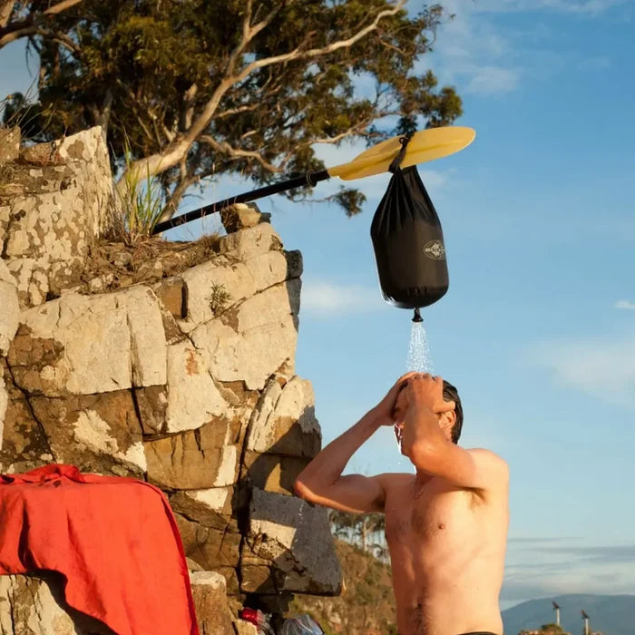 Person using a Sea To Summit Collapsible Pocket Shower on a rocky cliffside with a kayak paddle and towel in the background.
