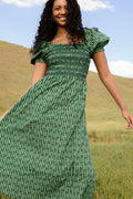 A woman stands outdoors in a grassy field, smiling in the Ivy City Co Sunday Best Dress in Pine Felicity Floral, with blue sky and hills behind her.