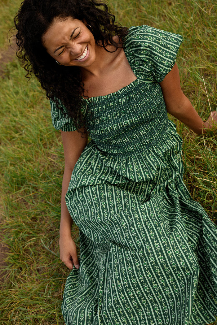 A woman with curly hair sits on grass, smiling with her eyes closed. She wears the Ivy City Co Sunday Best Dress in Pine Felicity Floral, featuring short puff sleeves and a smocked bodice.