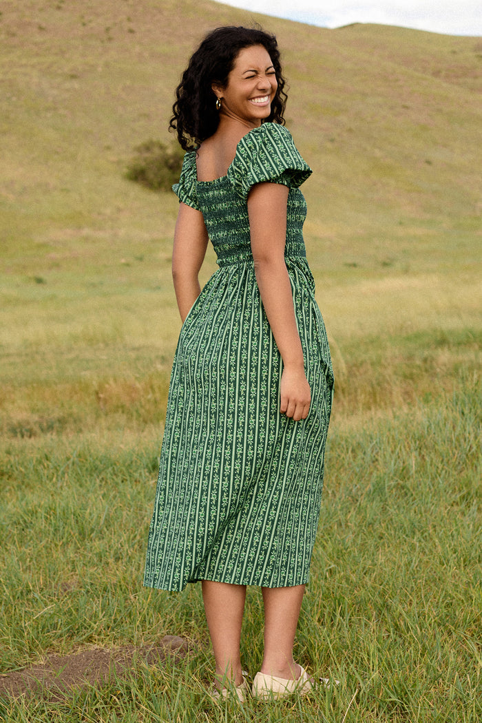 A woman with curly hair smiles outdoors on green grass, wearing the Ivy City Co Sunday Best Dress in Pine Felicity Floral with short sleeves and white shoes. Rolling grassy hills and a cloudy sky fill the background.