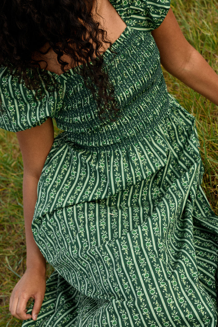 A person with curly hair sits on grass, wearing the Ivy City Co Sunday Best Dress in Pine Felicity Floral. The cropped photo highlights the dress’s short sleeves, floral stripe pattern, upper body, and a hand resting on the ground.