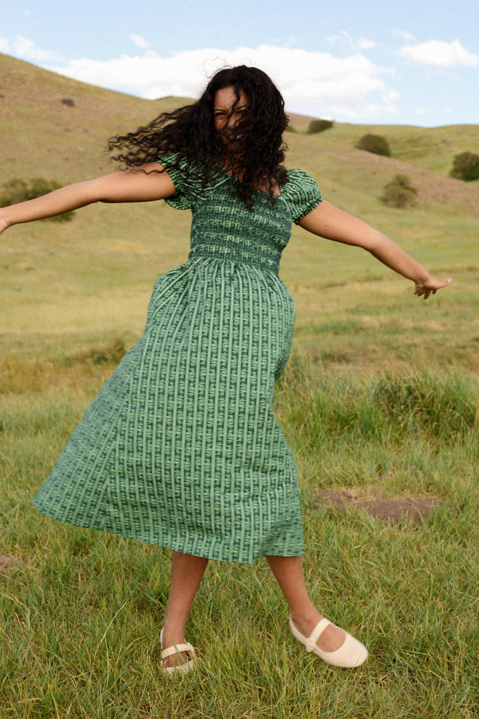 A woman twirls with outstretched arms in a grassy field beneath a partly cloudy sky, wearing Ivy City Co’s Sunday Best Dress in Pine Felicity Floral. Her curly hair flows as green hills rise in the background.