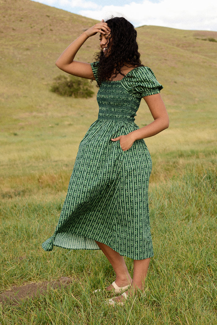 A woman wearing the Ivy City Co Sunday Best Dress in Pine Felicity Floral stands in a grassy field with hills behind her, holding her hand to her forehead and looking downward.