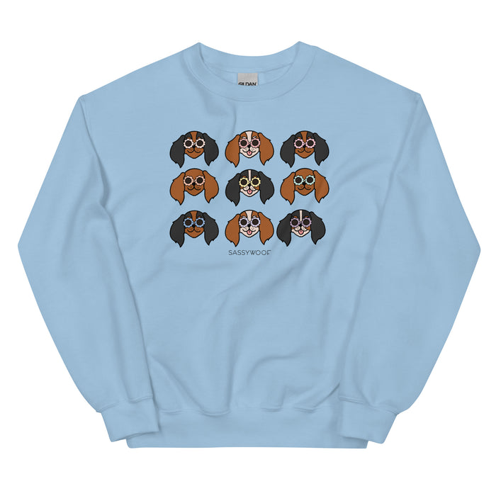The Sweatshirt - Cavaliers by Sassy Woof is a light blue unisex top featuring nine colorful cartoon dog faces in glasses and "SASSY WOOF" below—perfect for dog lovers who enjoy fun, playful style.