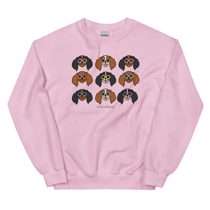 The "Sweatshirt - Cavaliers" by Sassy Woof is a pink unisex top with nine cartoon dog faces wearing glasses, each with unique fur designs. "SASSY WOOF" is printed below the playful illustrations—perfect for dog lovers!.
