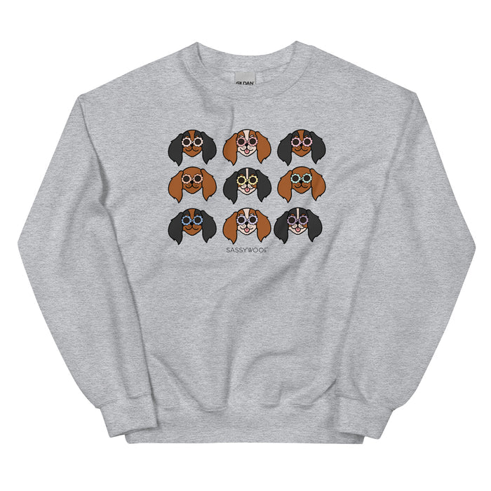 The Sassy Woof Sweatshirt - Cavaliers is a gray unisex sweatshirt with nine cartoon dog faces wearing round glasses in a grid, playful expressions, and SASSY WOOF printed below—perfect for dog lovers.
