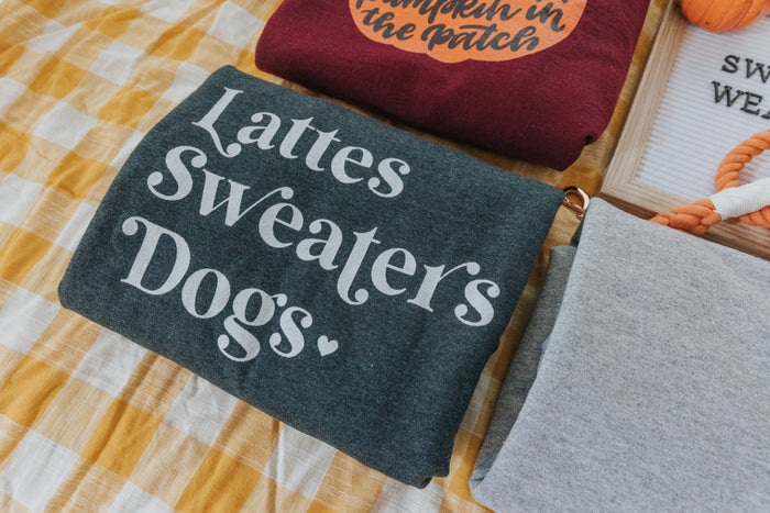 A folded dark gray Sassy Woof Sweatshirt - Lattes, Sweaters, Dogs sits on a yellow-and-white checkered surface beside other clothes and a letter board—an ideal unisex crewneck for cozy fall days.