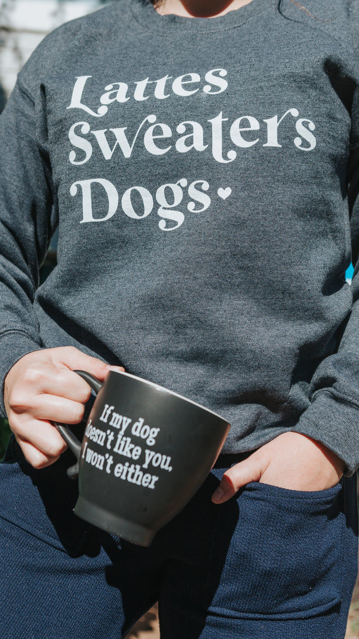 A person wears the Sassy Woof Sweatshirt - Lattes, Sweaters, Dogs and holds a black mug that says, “If my dog doesn’t like you, I won’t either.” This cozy unisex crewneck is perfect for fall and time with your pup.