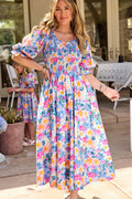 A woman wears the Ivy City Co Teagan Dress in Delia Blue—a colorful, floral midi with puff sleeves. Nearby, a young girl in a matching dress plays by patio furniture, creating a charming and maternity-friendly scene.
