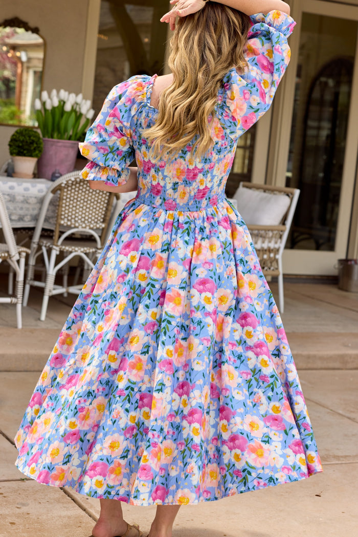A woman with long, wavy blonde hair stands barefoot outdoors in the Ivy City Co Teagan Dress in Delia Blue—a floral, maternity-friendly midi with puffed sleeves and a full skirt. She faces away, touching her hair amid patio furniture and flowers.