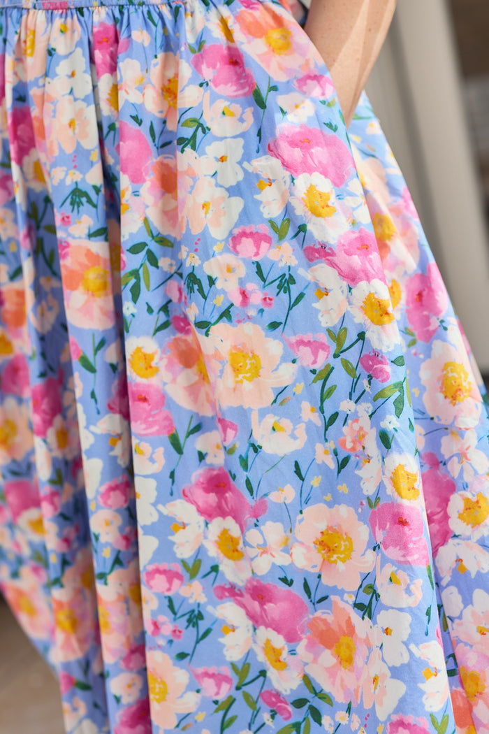 Close-up of the Teagan Dress in Delia Blue by Ivy City Co, a maternity-friendly blue floral midi dress with pink, yellow, and white flowers plus green leaves. A person’s arm rests near a pocket on the dress.
