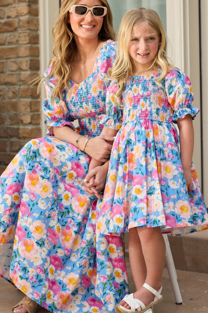 A woman and a young girl smile together in matching Ivy City Co Teagan Dress in Delia Blue, featuring blue, pink, and yellow florals. Both have long blonde hair; the woman wears sunglasses and the girl wears white sandals. Maternity friendly.