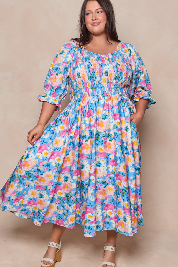 A woman smiles confidently in front of a neutral background wearing Ivy City Co’s Teagan Dress in Delia Blue, a vibrant floral midi with puffed sleeves and white sandals. This look is also maternity friendly.