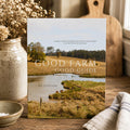 The book 'Good Farm Good Guide' 1st Edition standing upright on a rustic wooden table, surrounded by a ceramic jar of dried flowers, a small bowl, and a striped cloth. The cover features a pastoral landscape with a pond and grazing cows.