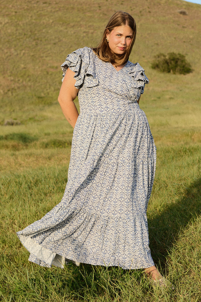 A woman with straight, shoulder-length hair stands in a grassy field wearing the Ivy City Co Trixie Dress in Blue Moroccan Tile, featuring ruffled sleeves. She faces the camera with gently rolling hills in the background.