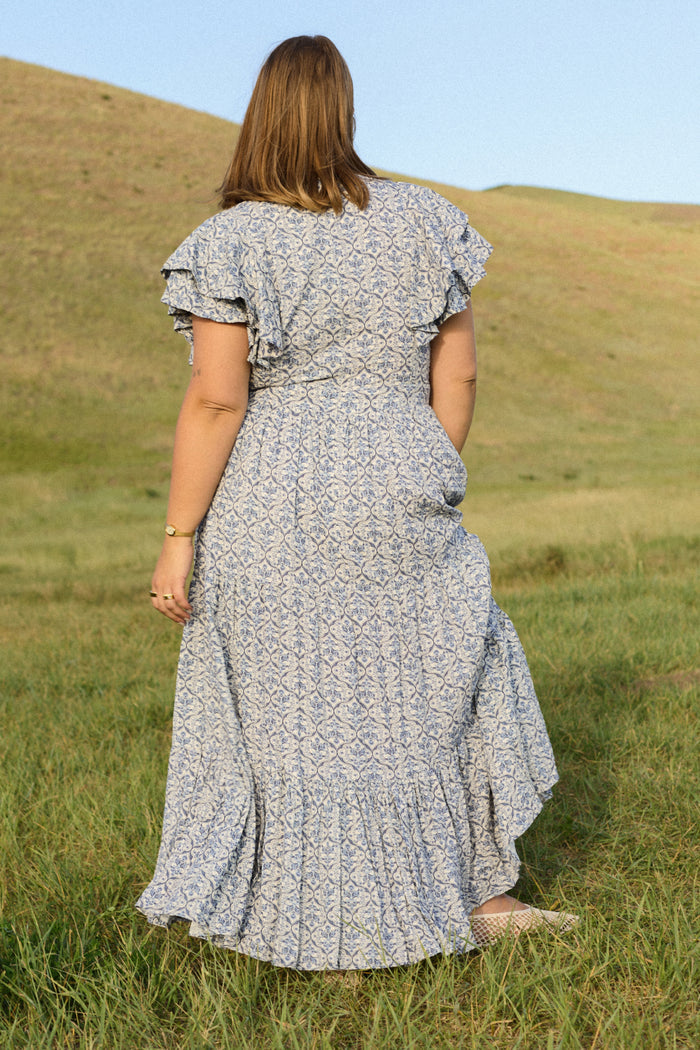 A woman with light brown hair stands in a grassy field, facing away, wearing the Trixie Dress in Blue Moroccan Tile by Ivy City Co. Rolling hills and a clear sky are visible in the background.