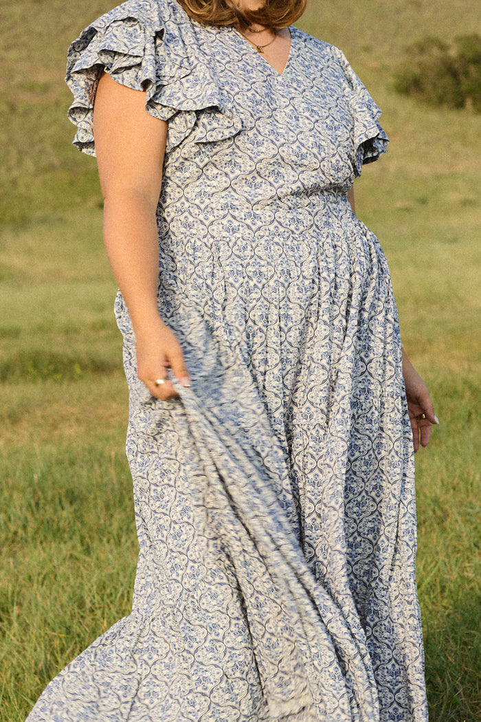 A person wears the Ivy City Co Trixie Dress in Blue Moroccan Tile with ruffled sleeves, standing on green grass and holding the skirt to the side. The photo is cropped at the shoulders and knees.