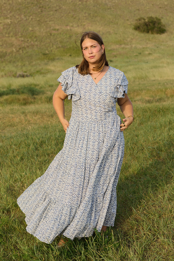A woman with shoulder-length brown hair and gold hoop earrings stands in a grassy field, wearing the Ivy City Co Trixie Dress in Blue Moroccan Tile, featuring short ruffled sleeves. She gazes directly at the camera.