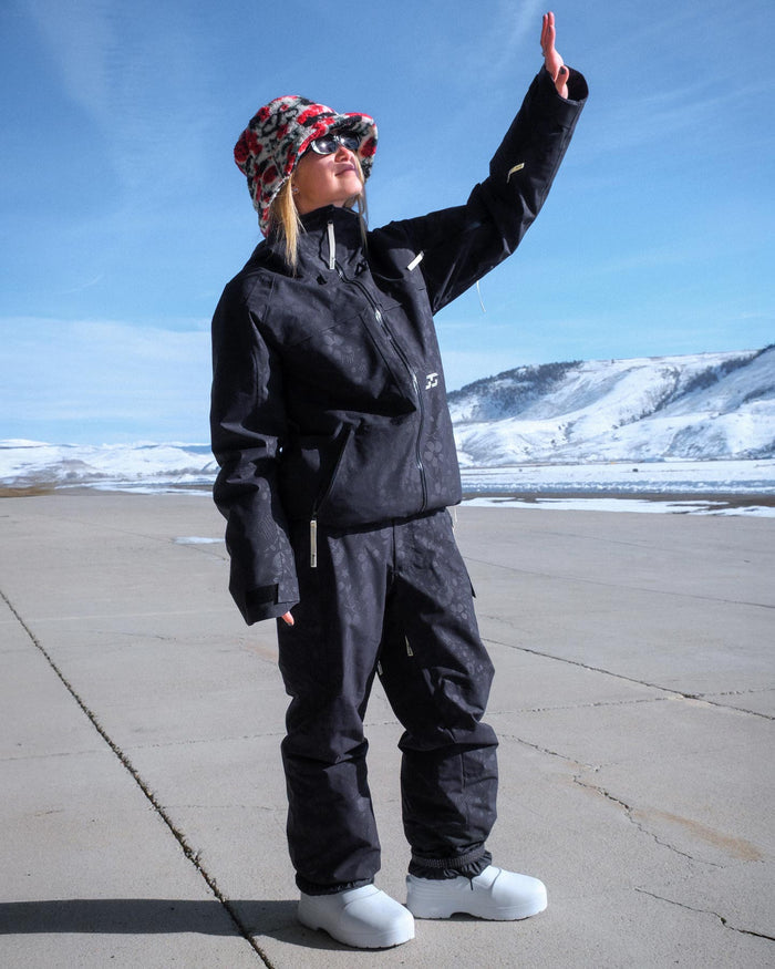 Person wearing a black snowsuit with a colorful beanie and sunglasses, standing on a snowy landscape.
