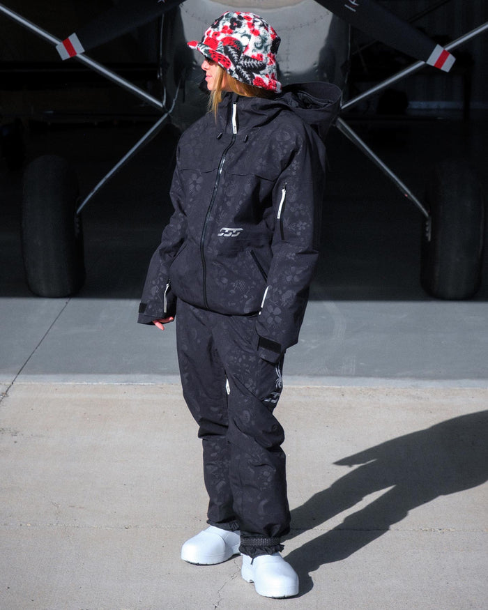 Person wearing a black snowsuit and colorful cap standing in front of an airplane.