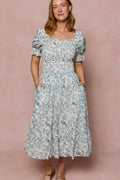 A woman with light brown hair stands against a light pink background, wearing Ivy City Co's Valencia Dress in Ivory Climbing Vine Bloom—a maternity-friendly floral midi dress with puff sleeves, square neckline, and pockets. She smiles slightly.