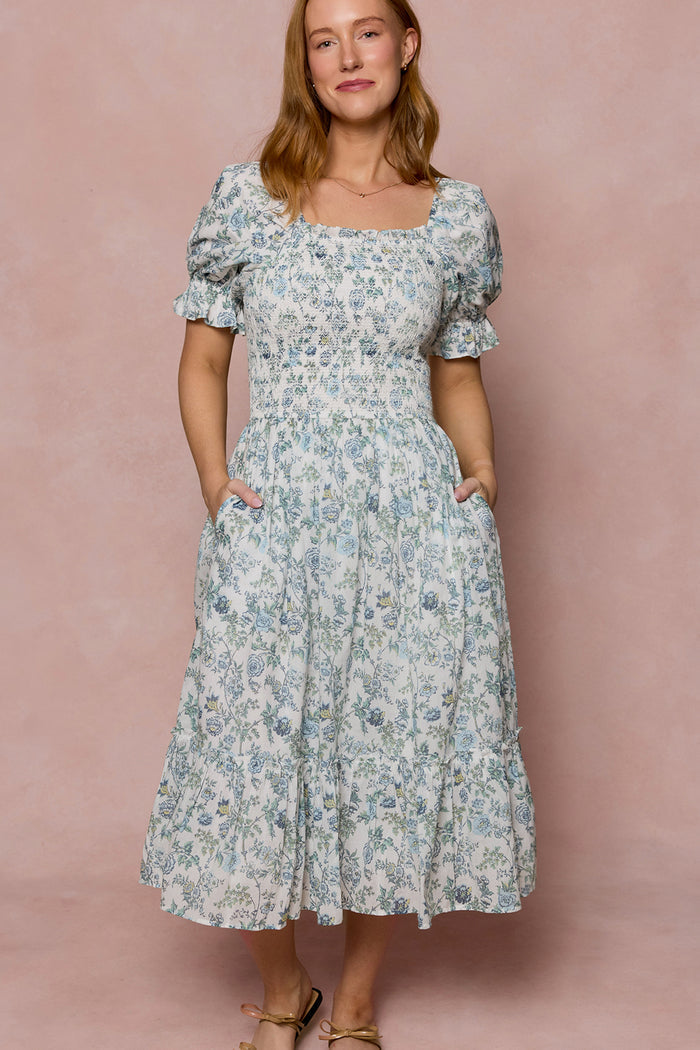 A woman with light brown hair stands against a light pink background, wearing Ivy City Co's Valencia Dress in Ivory Climbing Vine Bloom—a maternity-friendly floral midi dress with puff sleeves, square neckline, and pockets. She smiles slightly.