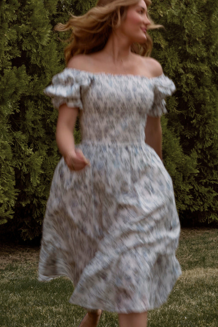 A woman runs on grass with greenery and trees behind her, hair flowing as she turns in the Ivy City Co Valencia Dress in Ivory Climbing Vine Bloom—an off-shoulder, maternity-friendly style that’s effortlessly elegant.