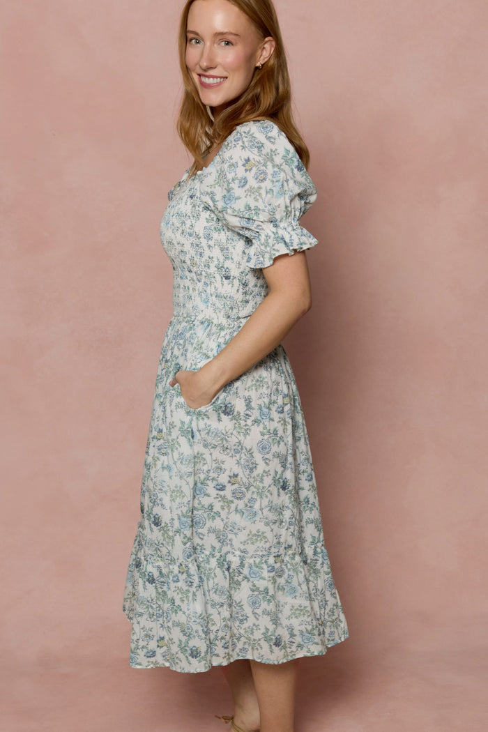A woman stands sideways, smiling, wearing the Ivy City Co Valencia Dress in Ivory Climbing Vine Bloom with puffed short sleeves and a ruffled hem, against a soft pink background.