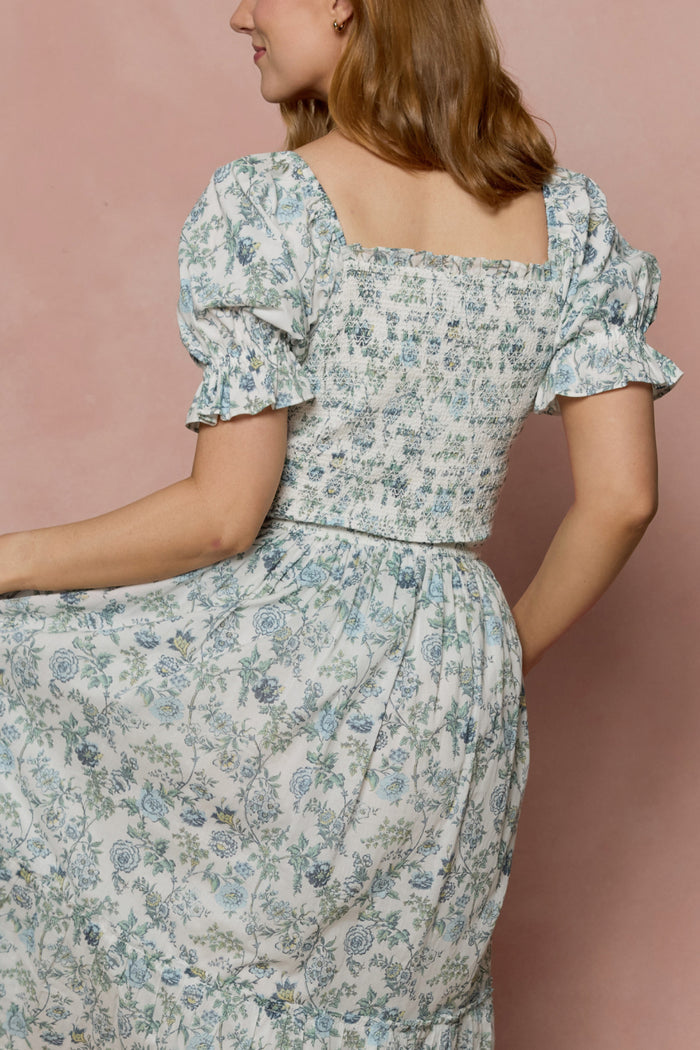 A woman with light brown hair models the Ivy City Co Valencia Dress in Ivory Climbing Vine Bloom. Featuring puffed short sleeves and a smocked, nursing-friendly bodice, she poses sideways holding her skirt against a pink backdrop.