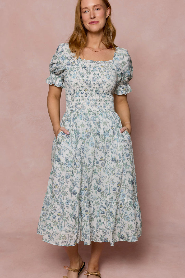 A woman in the Valencia Dress in Ivory Climbing Vine Bloom by Ivy City Co stands against a pink background, smiling with her hands in her pockets. This maternity-friendly dress combines comfort and style for every occasion.