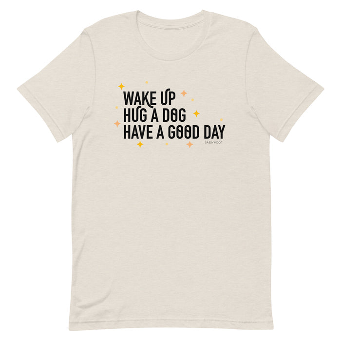 The Sassy Woof "Wake Up, Hug A Dog, Have A Good Day Tee" is a light beige unisex 100% cotton shirt featuring bold black text and orange/yellow star accents for comfy, all-day wear.