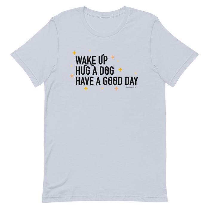 The Sassy Woof "Wake Up, Hug A Dog, Have A Good Day Tee" is a light gray unisex 100% cotton shirt featuring bold black text and small yellow stars for a comfy, cheerful style.