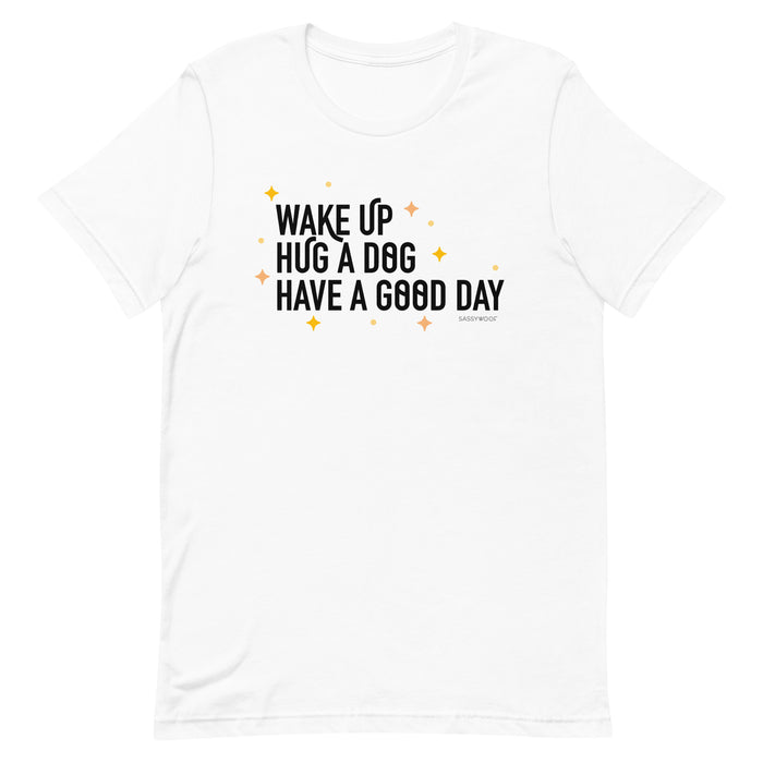 The Sassy Woof "Wake Up, Hug A Dog, Have A Good Day Tee" is a white unisex 100% cotton shirt featuring black text and small yellow star accents.