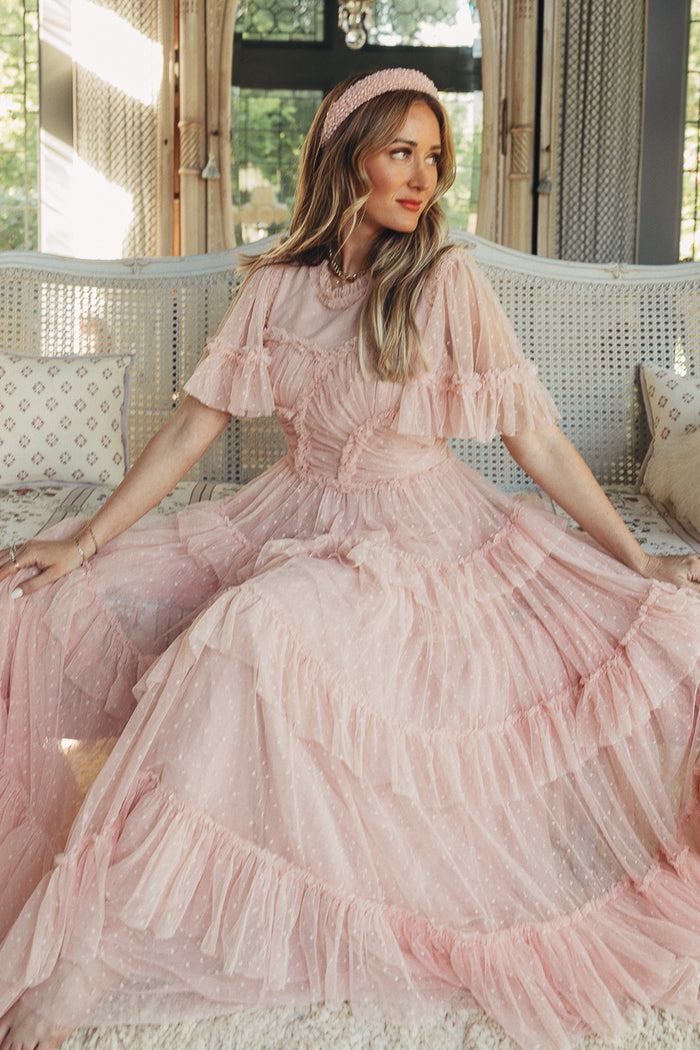 A woman in the Whimsical Dress in Blush by Ivy City Co, featuring sheer sleeves, sits gracefully on a white bench indoors. She wears a matching headband and gazes to the side, bathed in soft natural light.