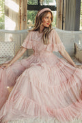 A woman in the Whimsical Dress in Blush by Ivy City Co, featuring sheer sleeves, sits gracefully on a white bench indoors. She wears a matching headband and gazes to the side, bathed in soft natural light.