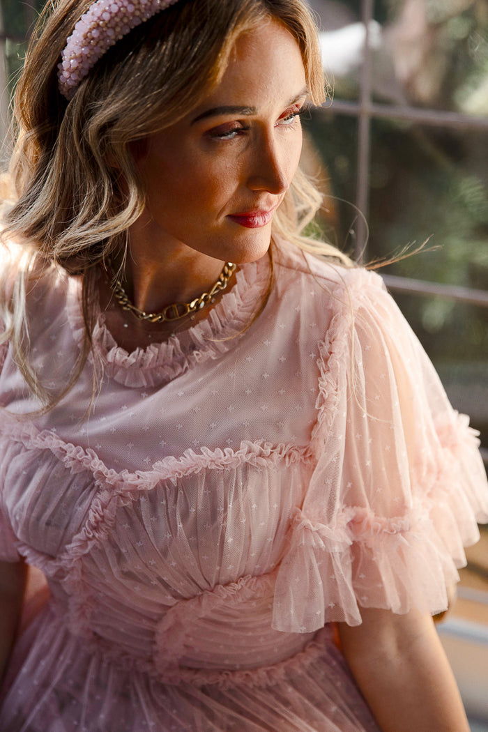 A woman in Ivy City Co’s Whimsical Dress in Blush, featuring sheer ruffles and puffed sleeves, pairs it with a textured headband and gold necklace. Sunlight highlights her wavy blonde hair as she gazes to the side.
